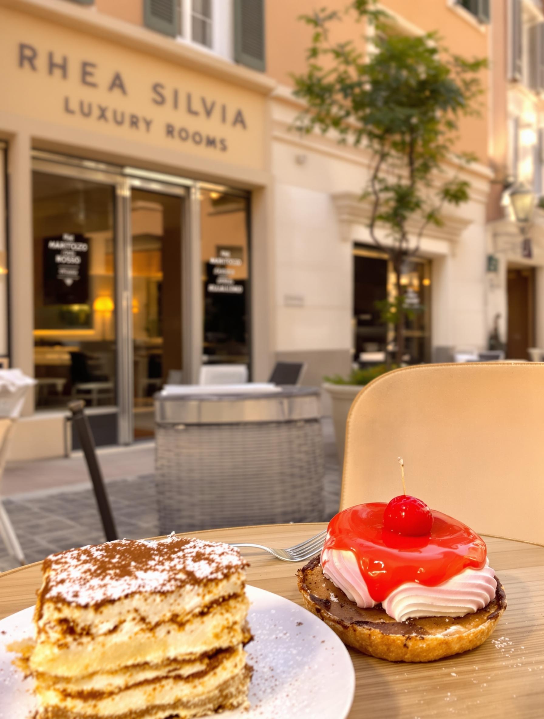 Table de dessert à Trastevere : tiramisu sur fond pastel, Maritozzo Rosso, lumière dorée, devanture Rhea Silvia Luxury Rooms en arrière-plan.